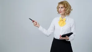Geschäftsfrau mit gelbem Halstuch zeigt mit einem Stift und hält ein Tablet in der anderen Hand vor grauem Hintergrund