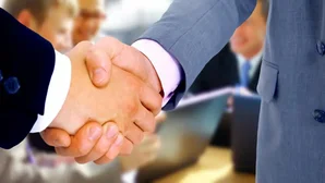 Zwei Geschäftsleute geben sich die Hand, im Hintergrund sitzen Personen in einem Besprechungsraum