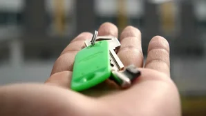 Hand hält einen Schlüsselbund mit grünem Anhänger vor unscharfem Hintergrund mit Reihenhäusern